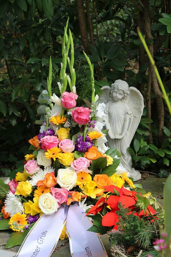 Blumenbouquet auf einer Grabstätte, im Hintergrund leicht unscharf angedeutet ein Grabengel, auf dem Spruchband in Deutsch "letzter Gruß"